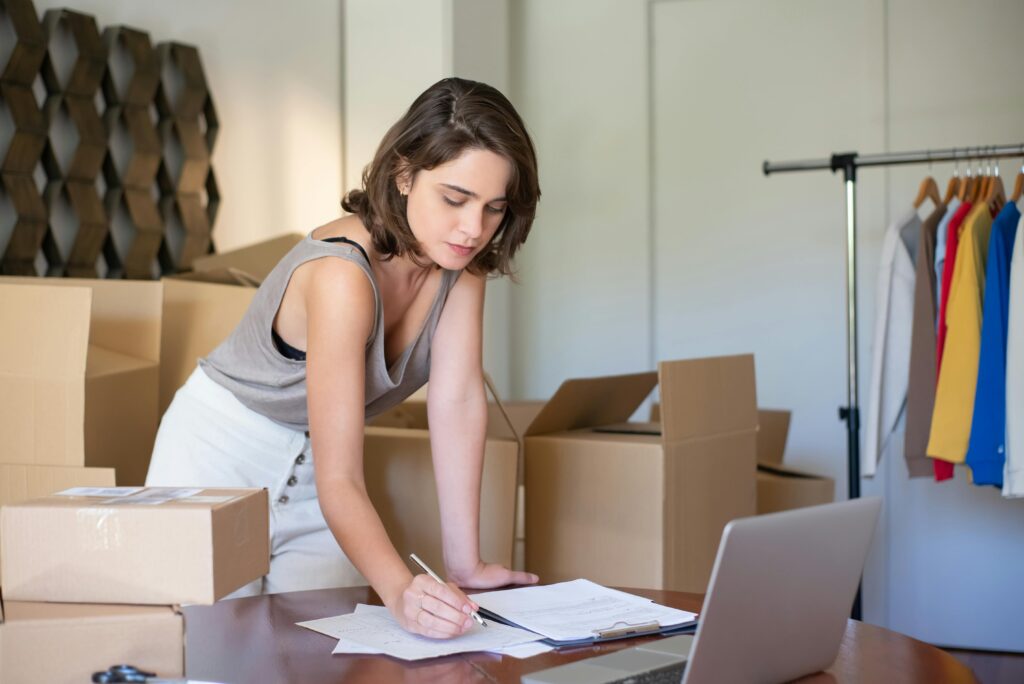 Emprendedora gestionando pedidos de su tienda online, rodeada de cajas y trabajando frente a una laptop que recibio por performance max
